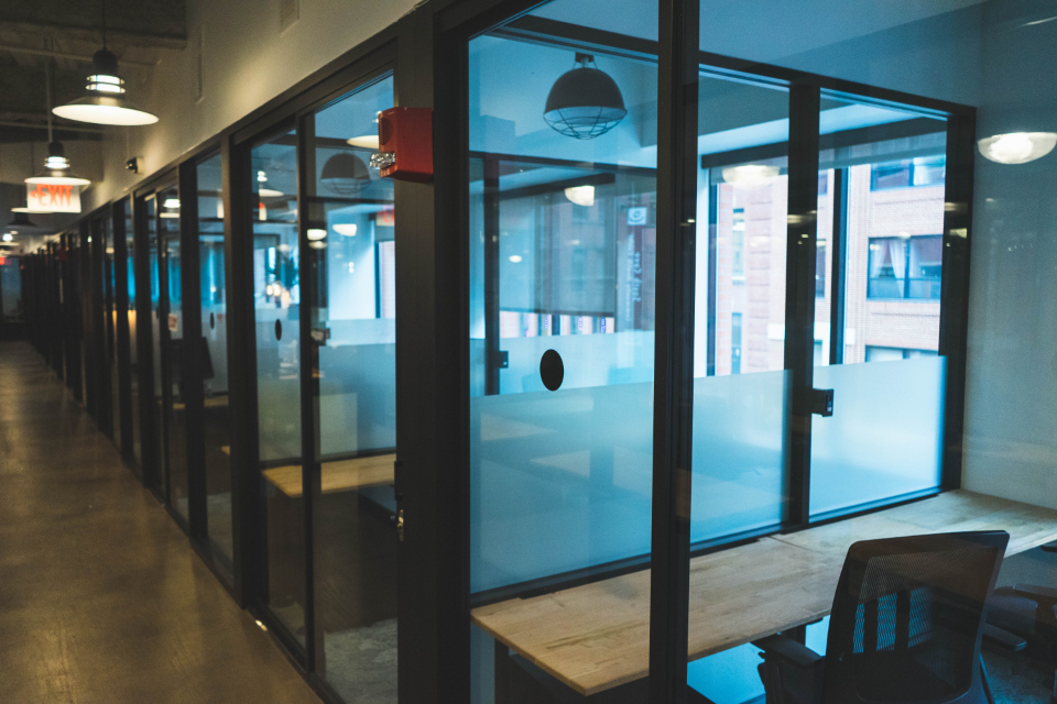 An empty modern office corridor lined with glass-walled rooms, softly lit in the evening